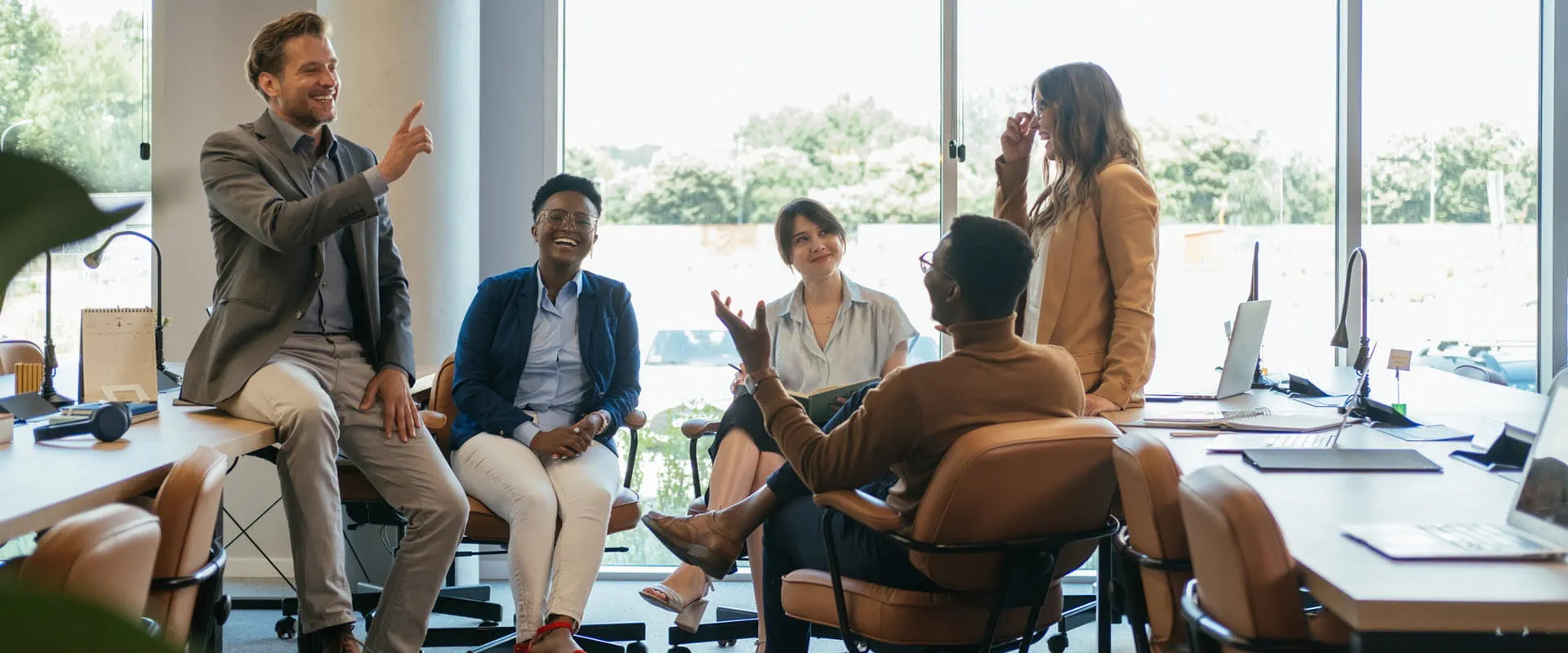 Five people sit and stand in a circle, laughing, in an office.