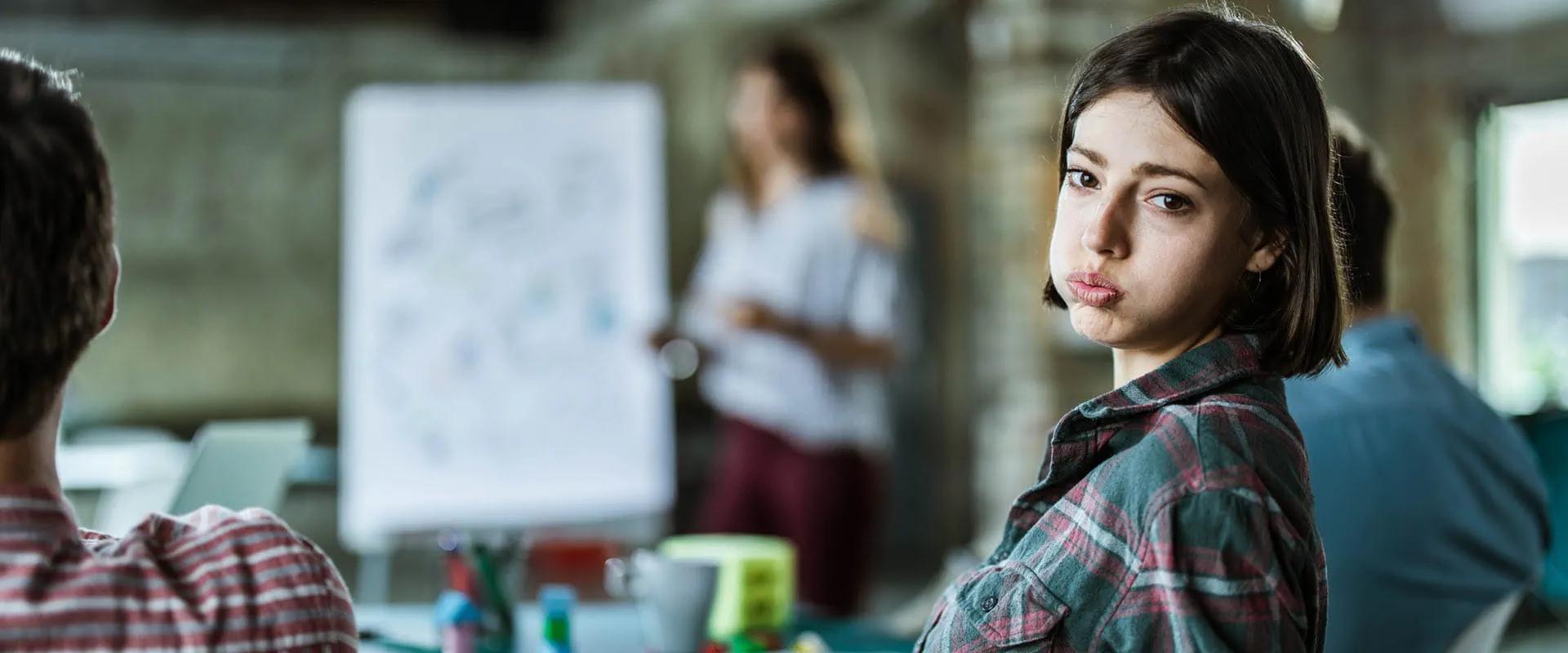 A young worker leans back in their chair, sighing and looking exasperated. In the background, their boss is leading a meeting. A young worker leans back in their chair, sighing and looking exasperated. In the background, their boss is leading a meeting.