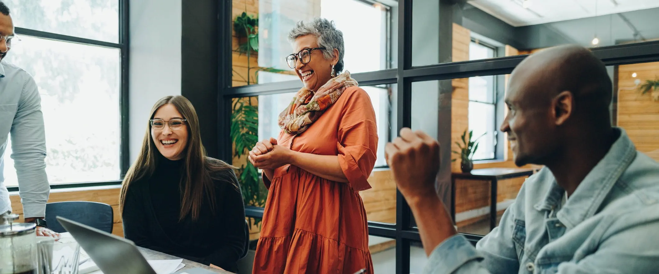 An laughing, older manager stand at an office conference table. Other workers are sitting and standing around the table laughing An laughing, older manager stand at an office conference table. Other workers are sitting and standing around the table laughing