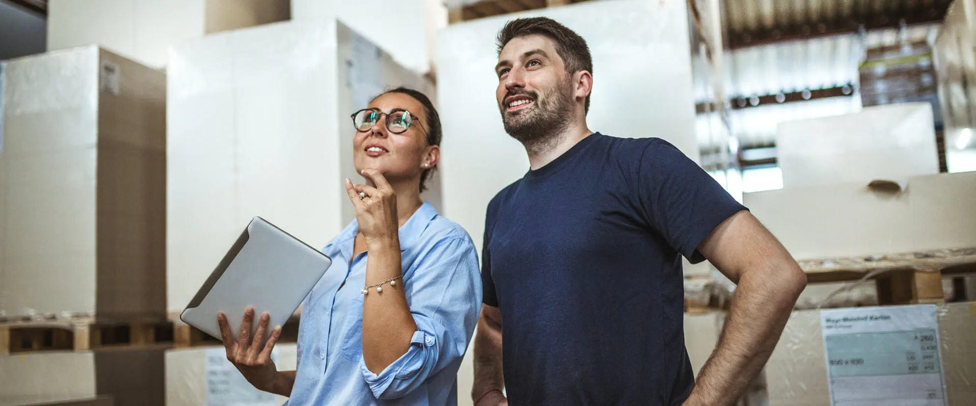 Two workers in a warehouse looking upward. One is holding a tablet. Two workers in a warehouse looking upward. One is holding a tablet.