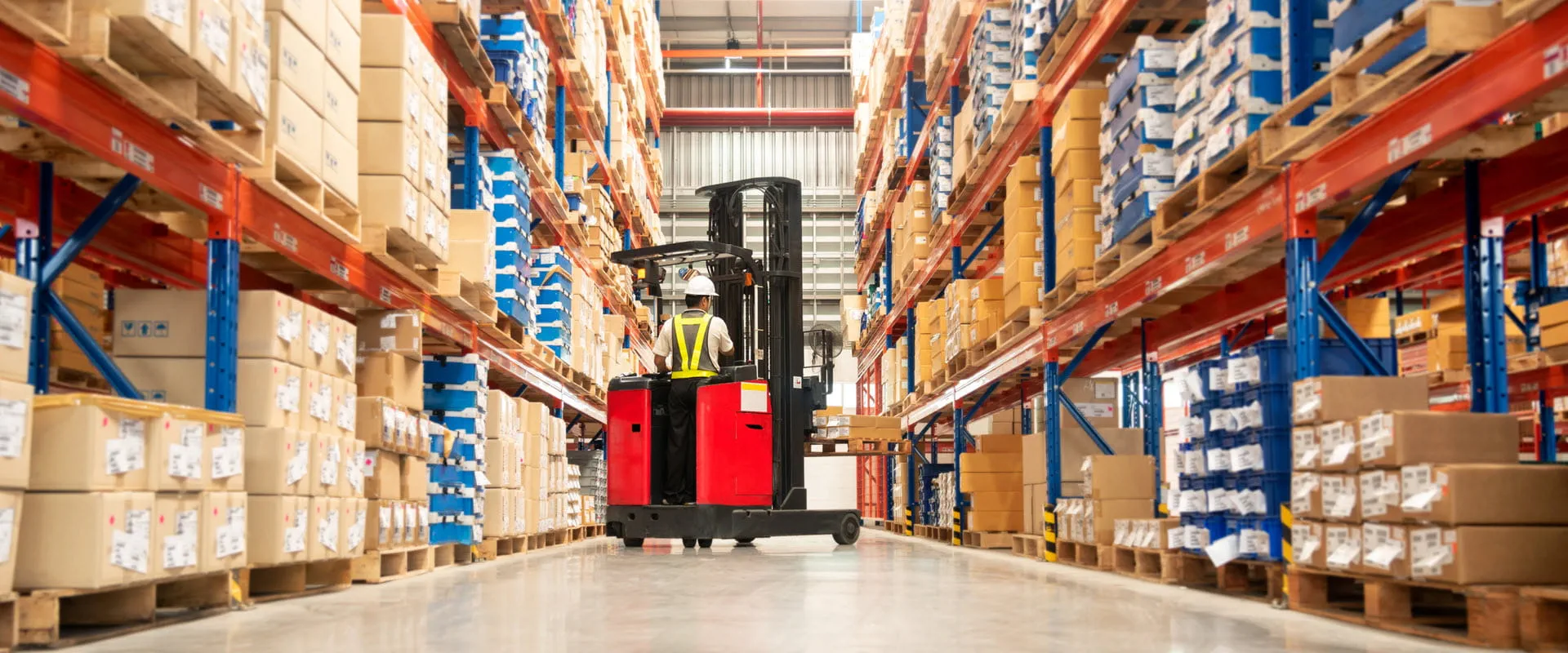 Worker in forklift-truck loading packed goods in huge distribution warehouse with high shelves.