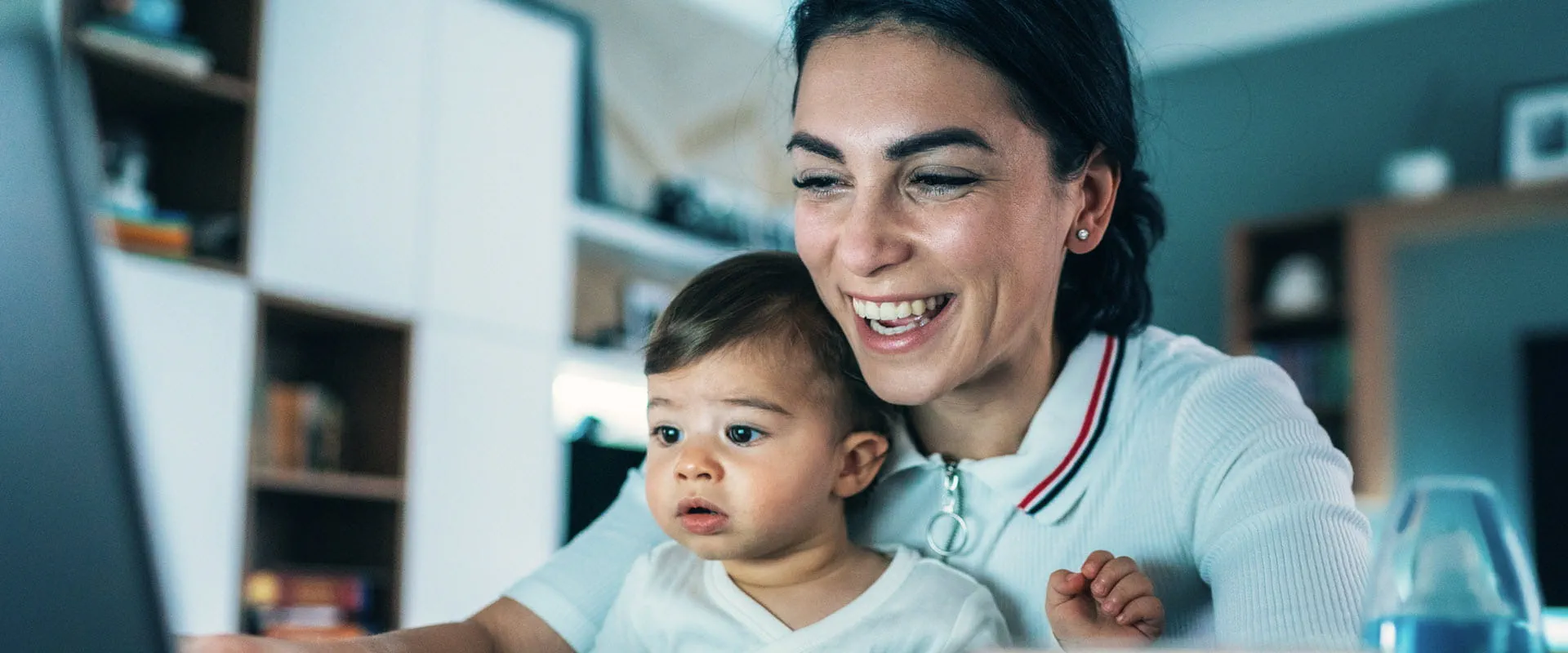A smiling parent at home, sits at their work laptop with a baby on their lap. A smiling parent at home, sits at their work laptop with a baby on their lap.