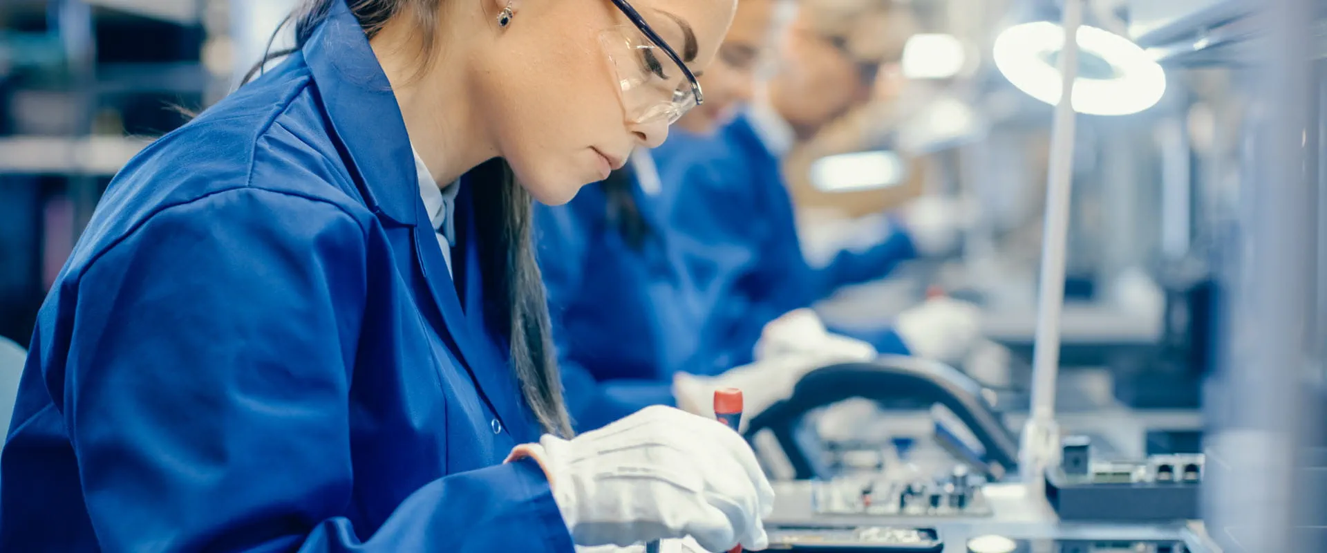 A worker dressed in blue coveralls, sits at a long table, inserting computer chips into devices. A worker dressed in blue coveralls, sits at a long table, inserting computer chips into devices.