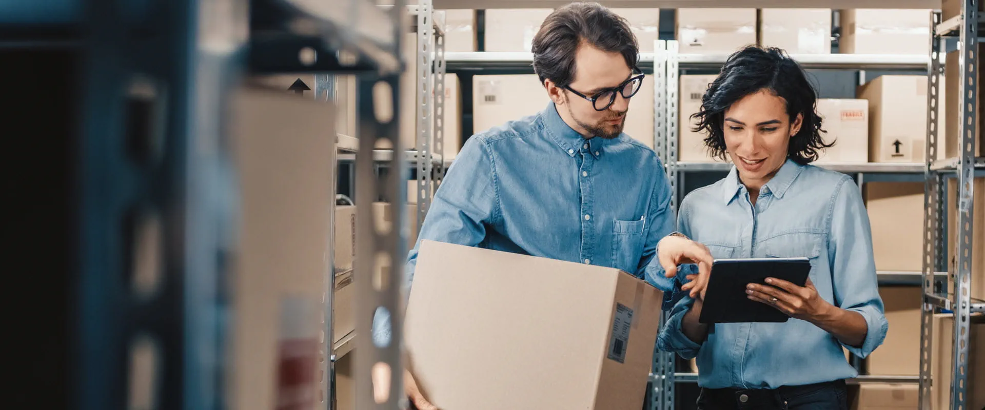 Two people stand in a warehouse, one holding a box, the other holding a tablet.