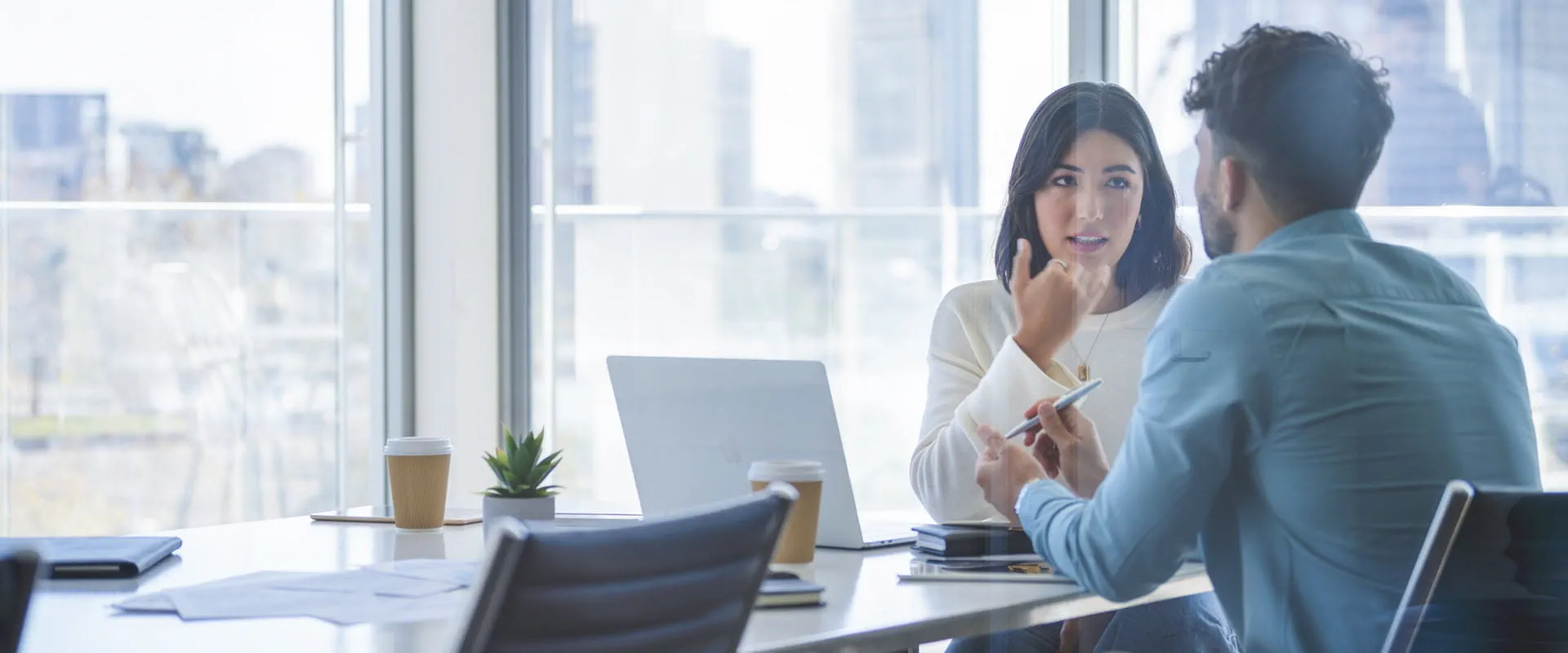 Two professionals having a focused discussion in a modern office with large windows and a city skyline in the background.