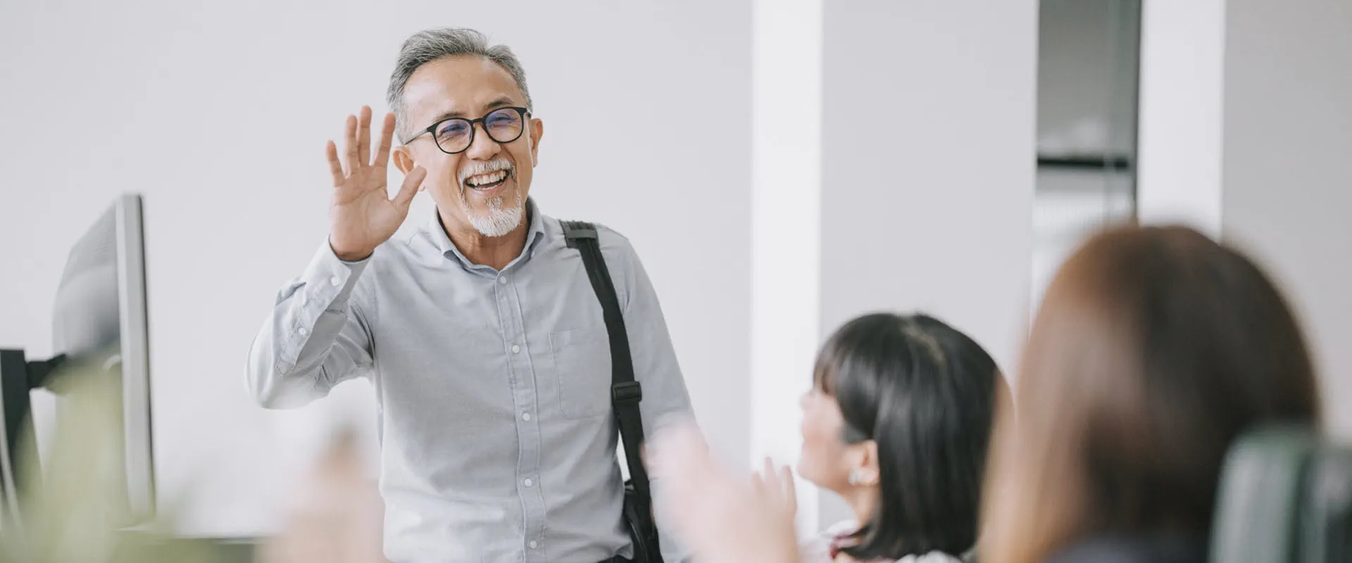A smiling person with glasses and a gray shirt waves as they enter a modern office space. A smiling person with glasses and a gray shirt waves as they enter a modern office space.