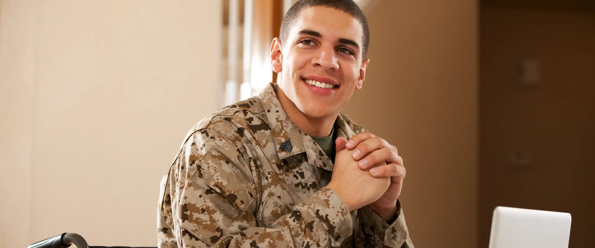 A smiling individual in military fatigues in a wheelchair sits at a table with a laptop on it. A smiling individual in military fatigues in a wheelchair sits at a table with a laptop on it.