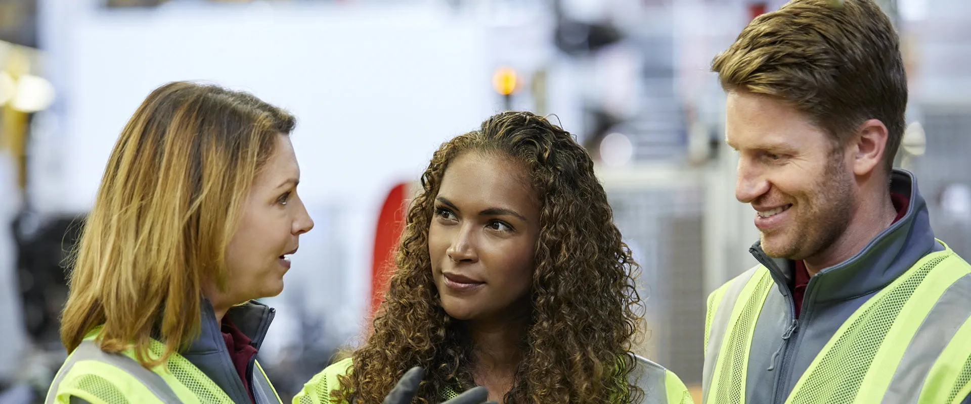 Three professionals wearing high-visibility safety vests engaged in a discussion at a manufacturing facility. 