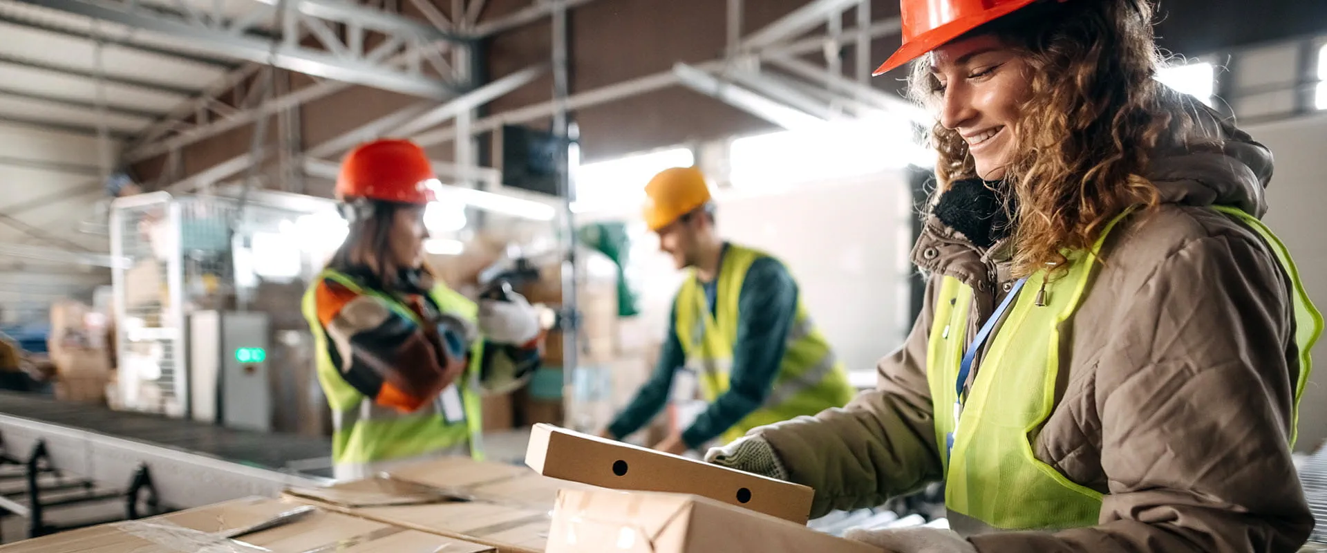 Three workers in a distribution center wearing hard hats and safety vests, sort packages. Three workers in a distribution center wearing hard hats and safety vests, sort packages.