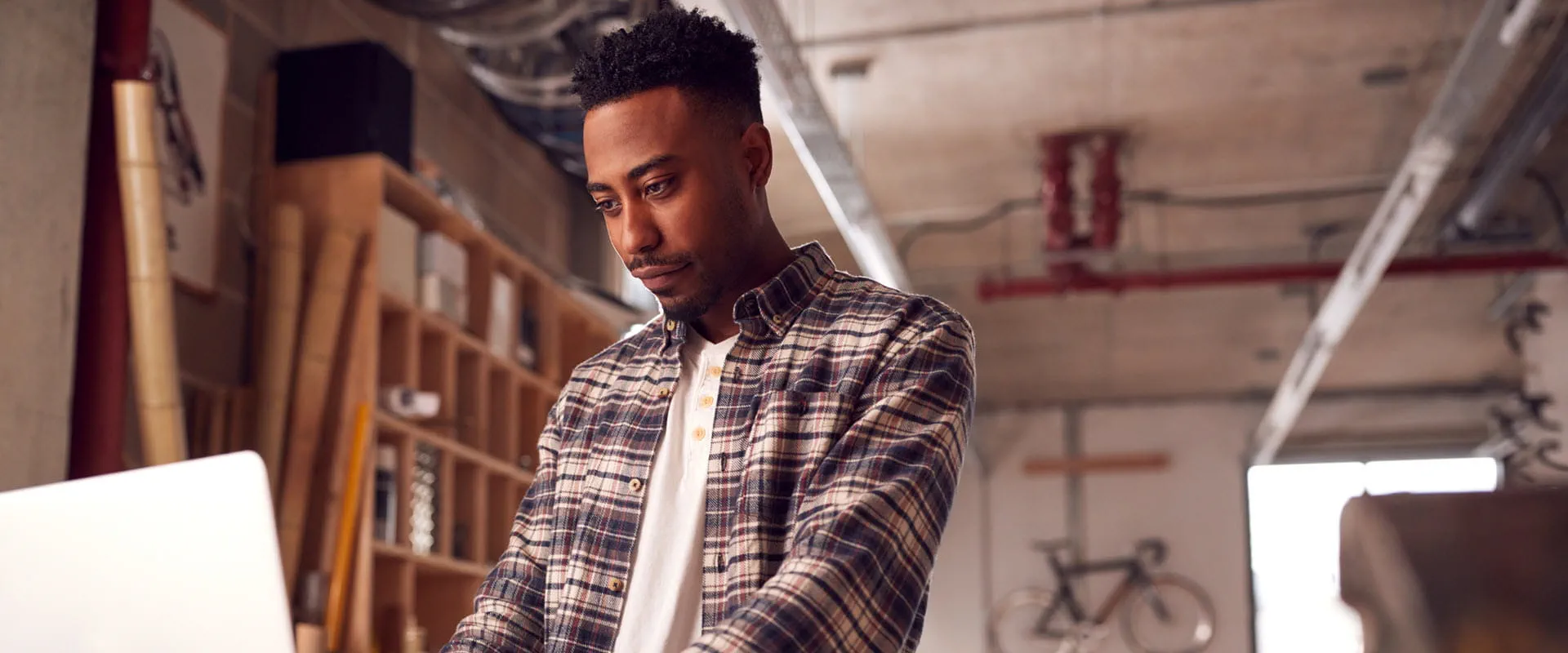 A focused individual wearing a plaid shirt works on a laptop in a creative workspace with industrial-style decor.