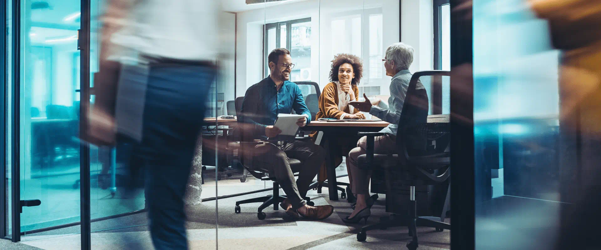 A group of three people sit in a conference room and talk, while in the middle of a busy office