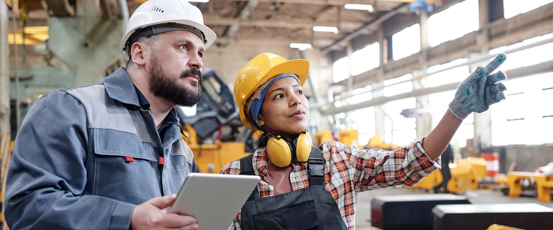 Two workers in hard hats and coveralls look at something in the distance in a manufacturing warehouse. Two workers in hard hats and coveralls look at something in the distance in a manufacturing warehouse.