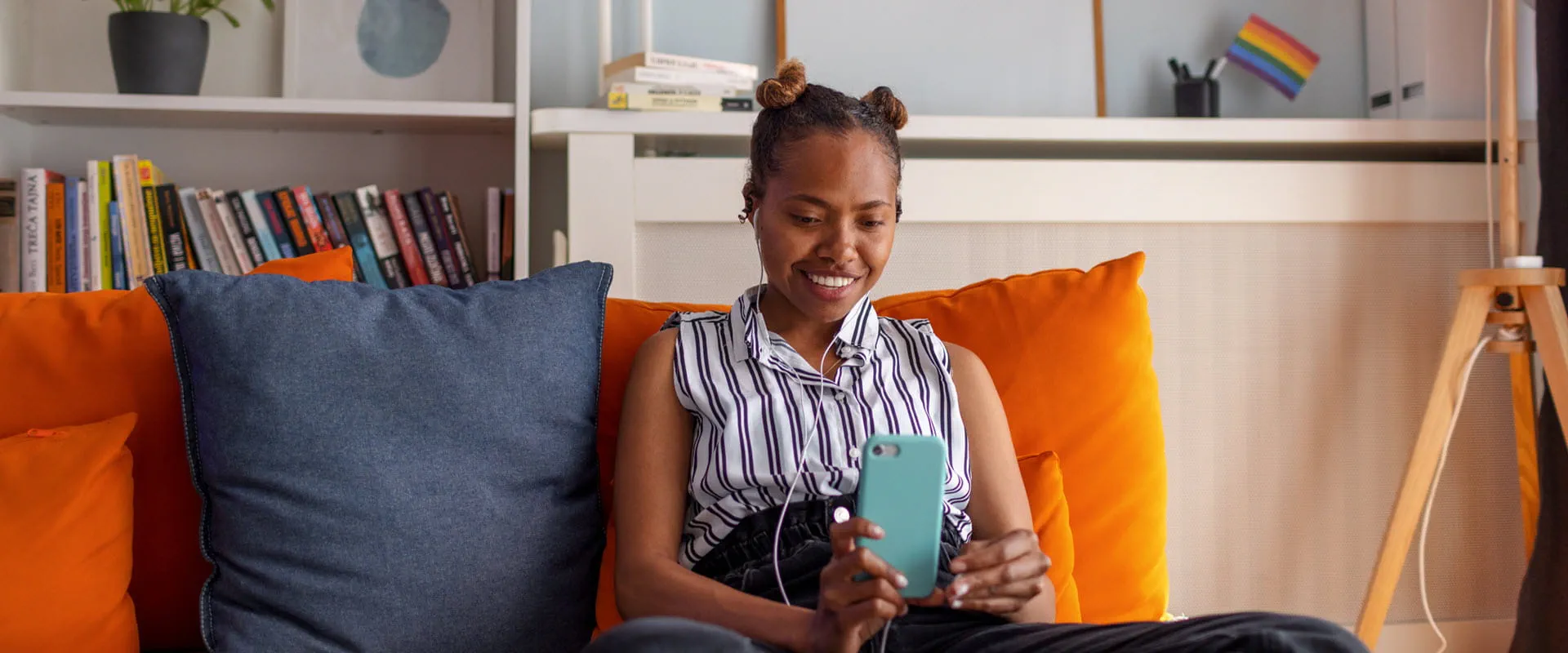 A fashionably dressed young person sits on an orange couch looking at their phone with headphones in A fashionably dressed young person sits on an orange couch looking at their phone with headphones in