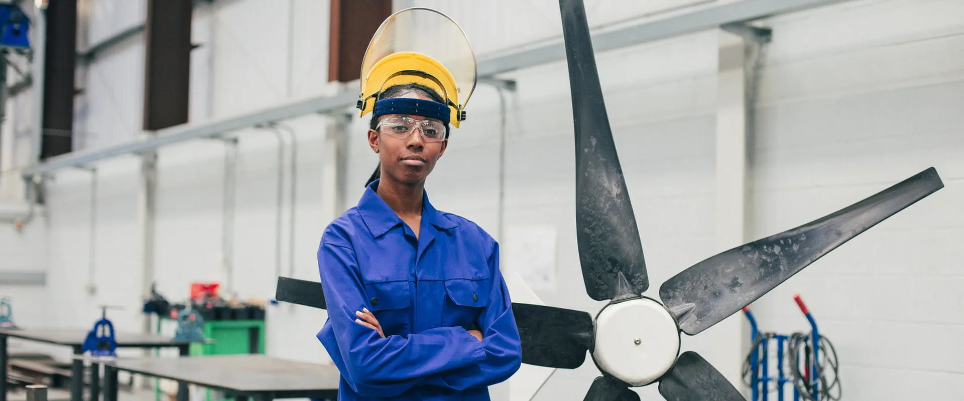 A woman in blue coveralls, safety glasses, and a hard hat, stands in front of an unfinished windmill propeller, in a workspace A woman in blue coveralls, safety glasses, and a hard hat, stands in front of an unfinished windmill propeller, in a workspace