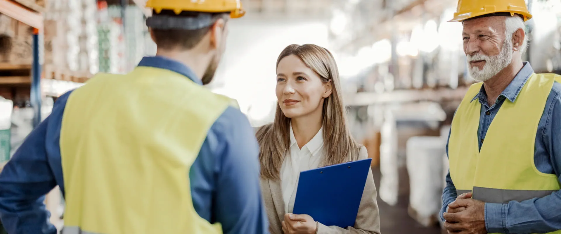 A person in a warehouse shakes hands with someone in a hard hat and safety vest. Behind them stands an older worker also wearing a hard hat and safety vest.