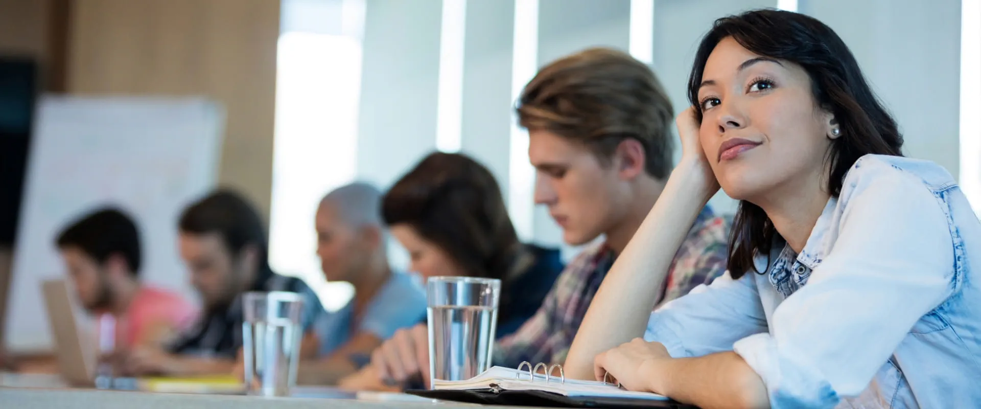 A young worker in a denim button-up, sits at the end of a crowded conference table, and seems to be day-dreaming off into the distrance