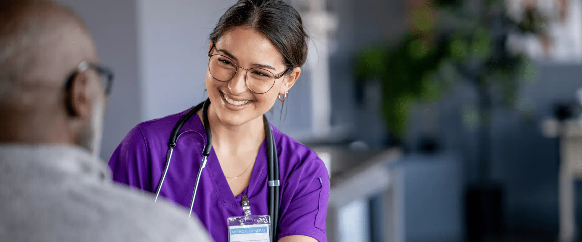 A young medical professional in scrubs smiles and talks to a patient.
