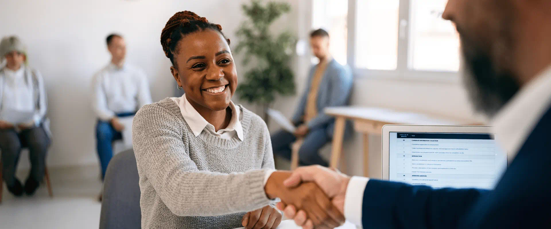 A smiling job candidate shakes hand with an interviewer. Behind them, others sit at chairs waiting to be interviewed. A smiling job candidate shakes hand with an interviewer. Behind them, others sit at chairs waiting to be interviewed.