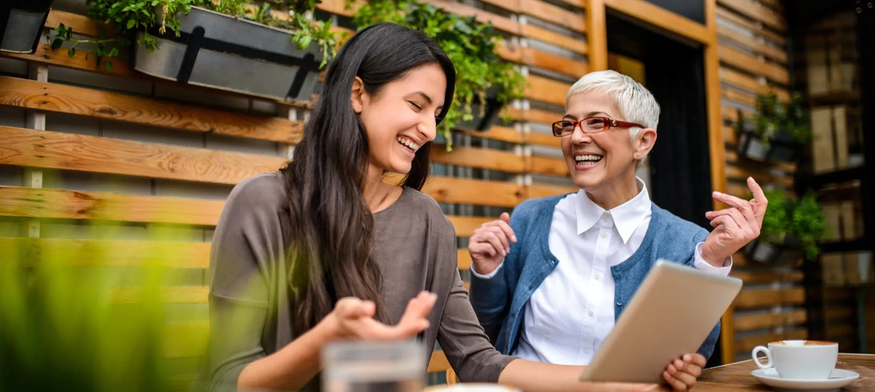 A younger and older coworker sit next to one another at a coffee shop. They are laughing. A younger and older coworker sit next to one another at a coffee shop. They are laughing.