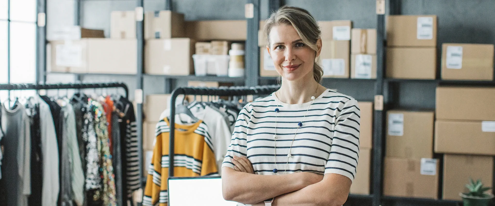 A business owner happily looks at the camera. Behind them are boxes stack on shelves, clothes on racks, and a laptop on a desk.