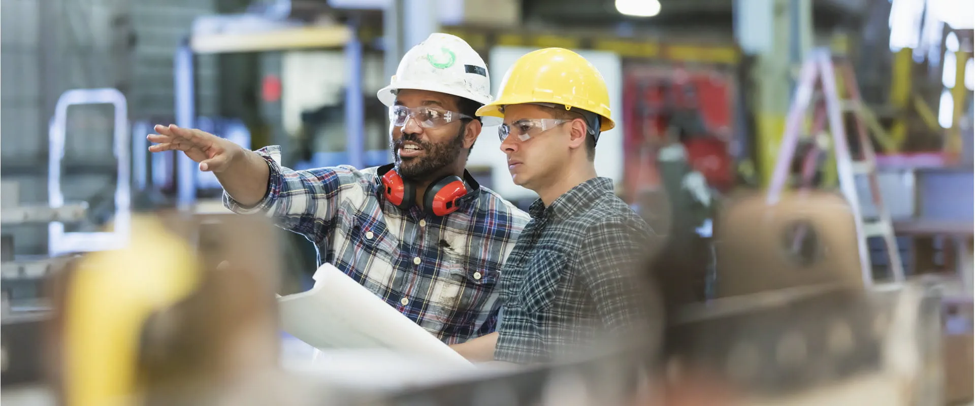Two industrial workers wearing hard hats and safety glasses stand in a manufacturing facility. Two industrial workers wearing hard hats and safety glasses stand in a manufacturing facility.