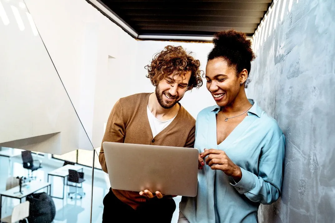 Two professionals standing in a modern office hallway reviewing information together on a laptop.