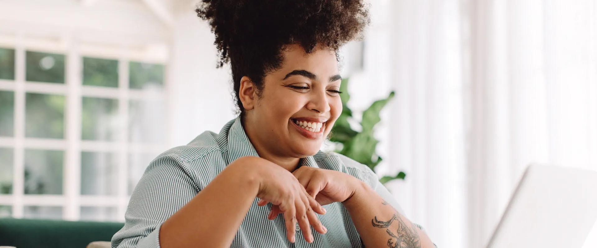 A smiling individual with curly hair and tattoos works on a laptop in a bright, home-like setting. A smiling individual with curly hair and tattoos works on a laptop in a bright, home-like setting.