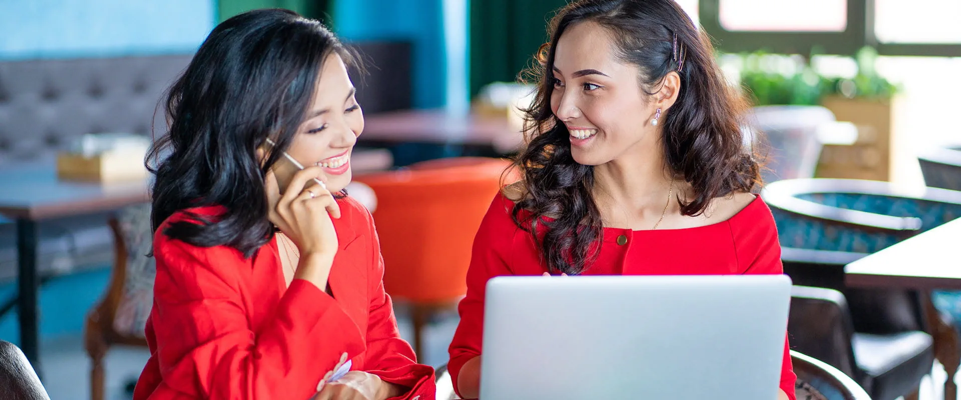 Two women, both wearing red, sitting at table working on a laptop. One is using a cell phone.
