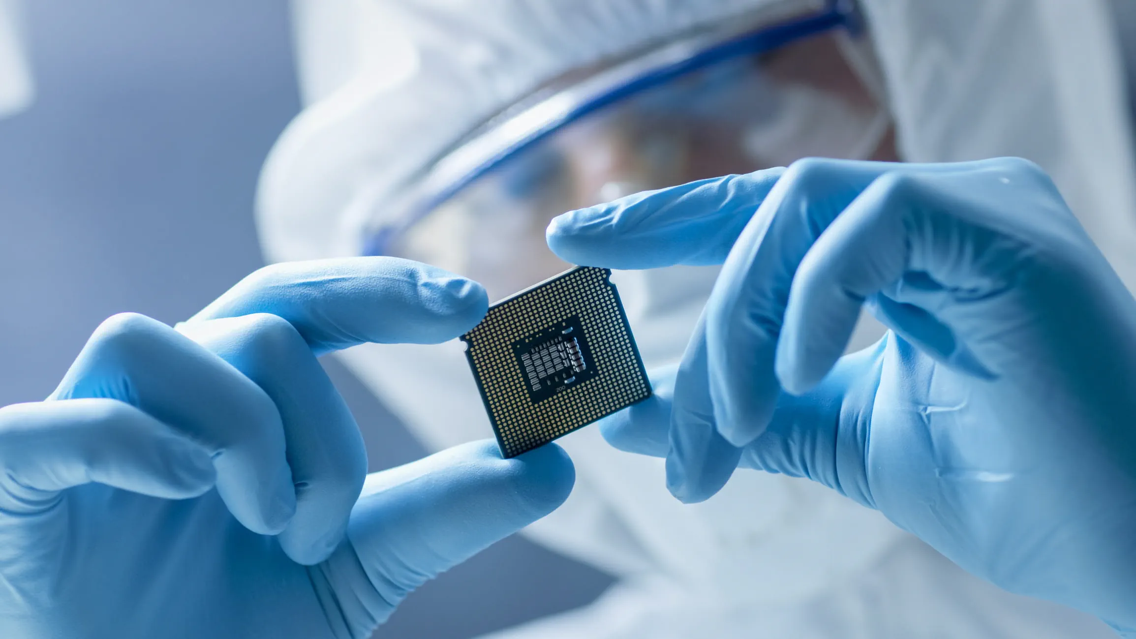A technician wearing blue gloves and protective gear carefully holding a microchip in a cleanroom environment. A technician wearing blue gloves and protective gear carefully holding a microchip in a cleanroom environment.