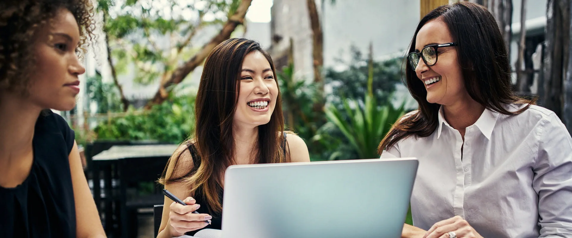 A group of people talk, while sitting outside at a table with notebooks and a laptop