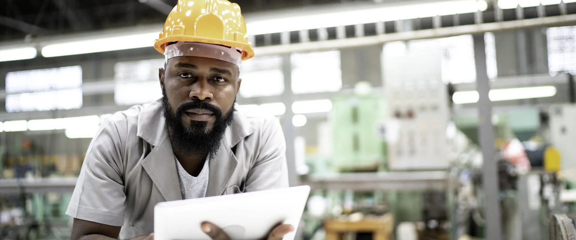 On a factory floor, an individual in coveralls and a hardhat, leans over a work table with a tablet in their hand