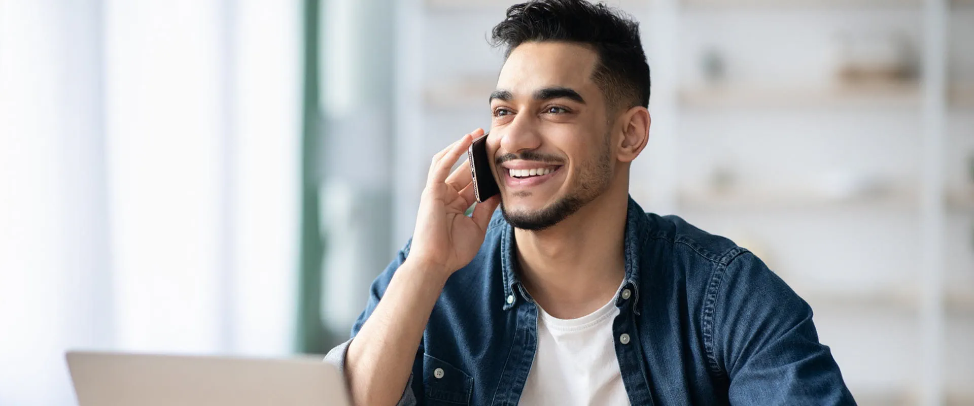 A smiling individual working from home, sitting at a desk with a laptop while talking on the phone. A smiling individual working from home, sitting at a desk with a laptop while talking on the phone.