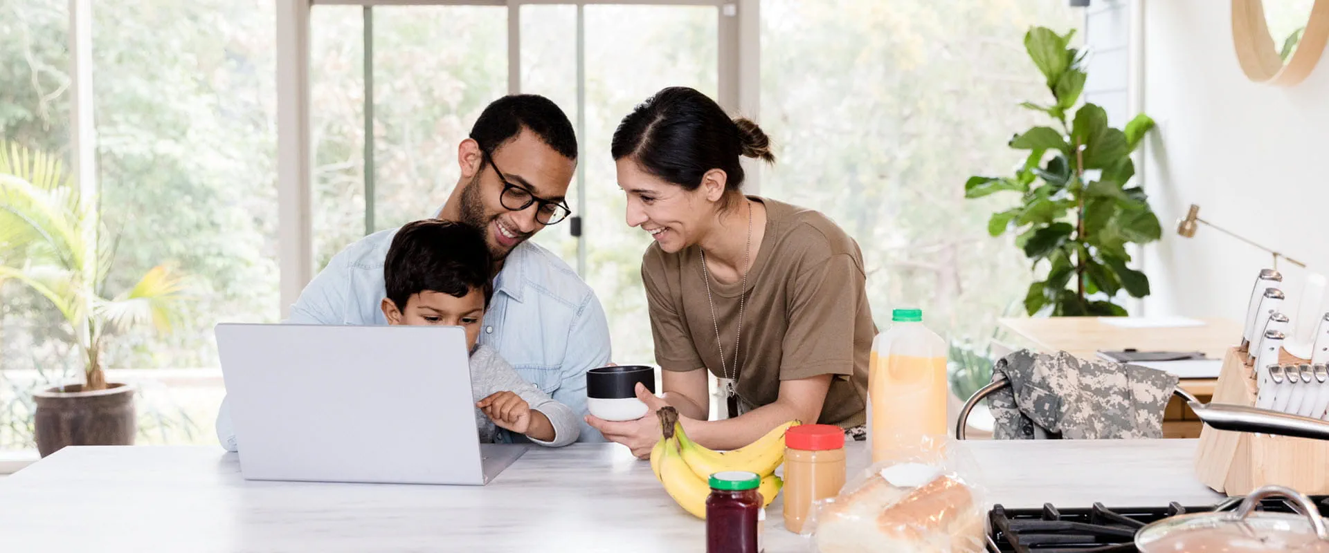 Two parents, one in a military uniform, sit at a kitchen island with their child in between them. There is a laptop and breakfast on the table. Two parents, one in a military uniform, sit at a kitchen island with their child in between them. There is a laptop and breakfast on the table.