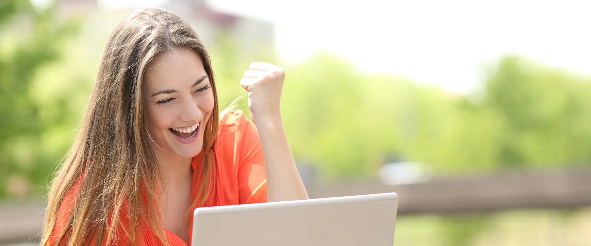 A person sitting outdoors, wearing an orange blouse, smiling and raising a fist in excitement while looking at a laptop screen. A person sitting outdoors, wearing an orange blouse, smiling and raising a fist in excitement while looking at a laptop screen.