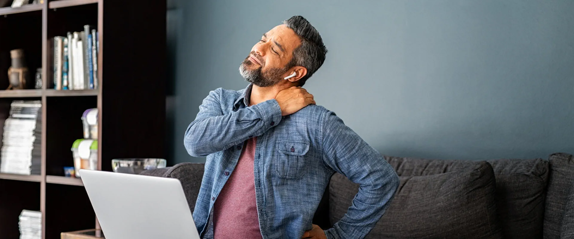A middle-aged worker sits on an office sofa with a laptop on his knees. He is stretching his back as if he's uncomfortable.