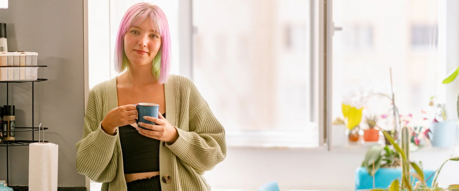 A young person, with pink and green dyed hair, in a crop top and cardigan leans against the wall and holds a blue mug, while smiling. A young person, with pink and green dyed hair, in a crop top and cardigan leans against the wall and holds a blue mug, while smiling.