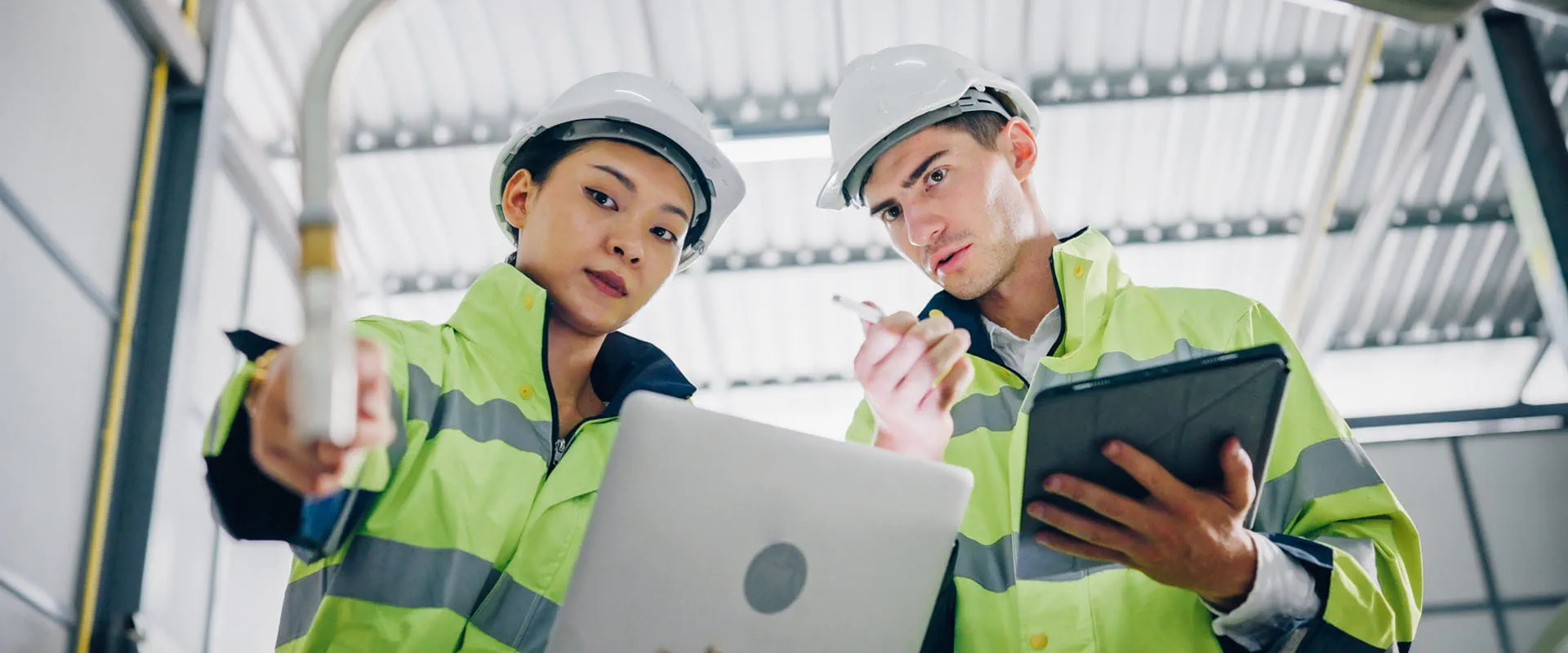 Two workers in safety jackets and hard hats look at a robotic arm in a manufacturing facility. Two workers in safety jackets and hard hats look at a robotic arm in a manufacturing facility.