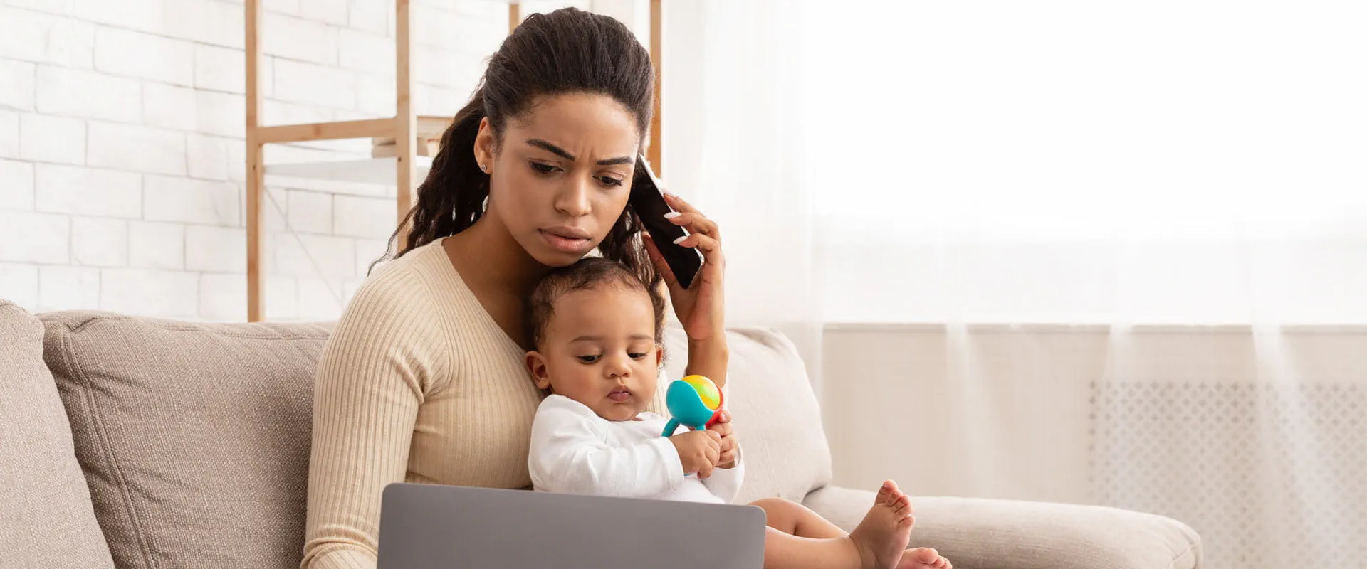 : A parent working from home sits on a couch with a laptop, holding a baby. : A parent working from home sits on a couch with a laptop, holding a baby.