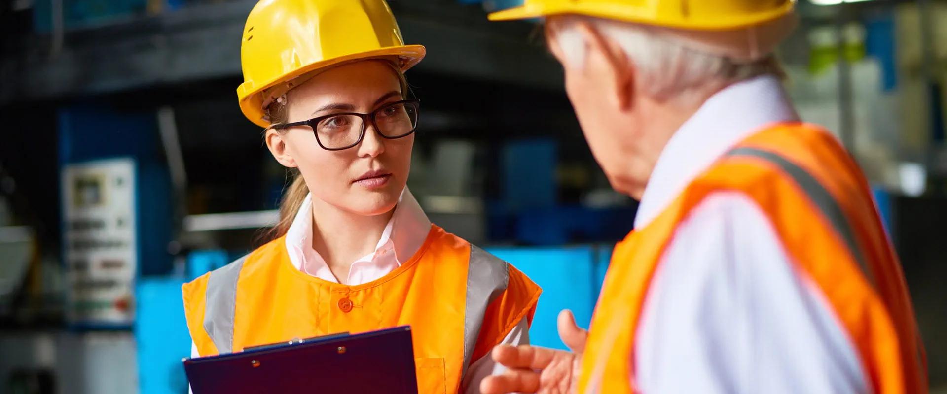 Two professionals in a manufacturing or industrial setting wearing high-visibility orange safety vests and yellow hard hats.
