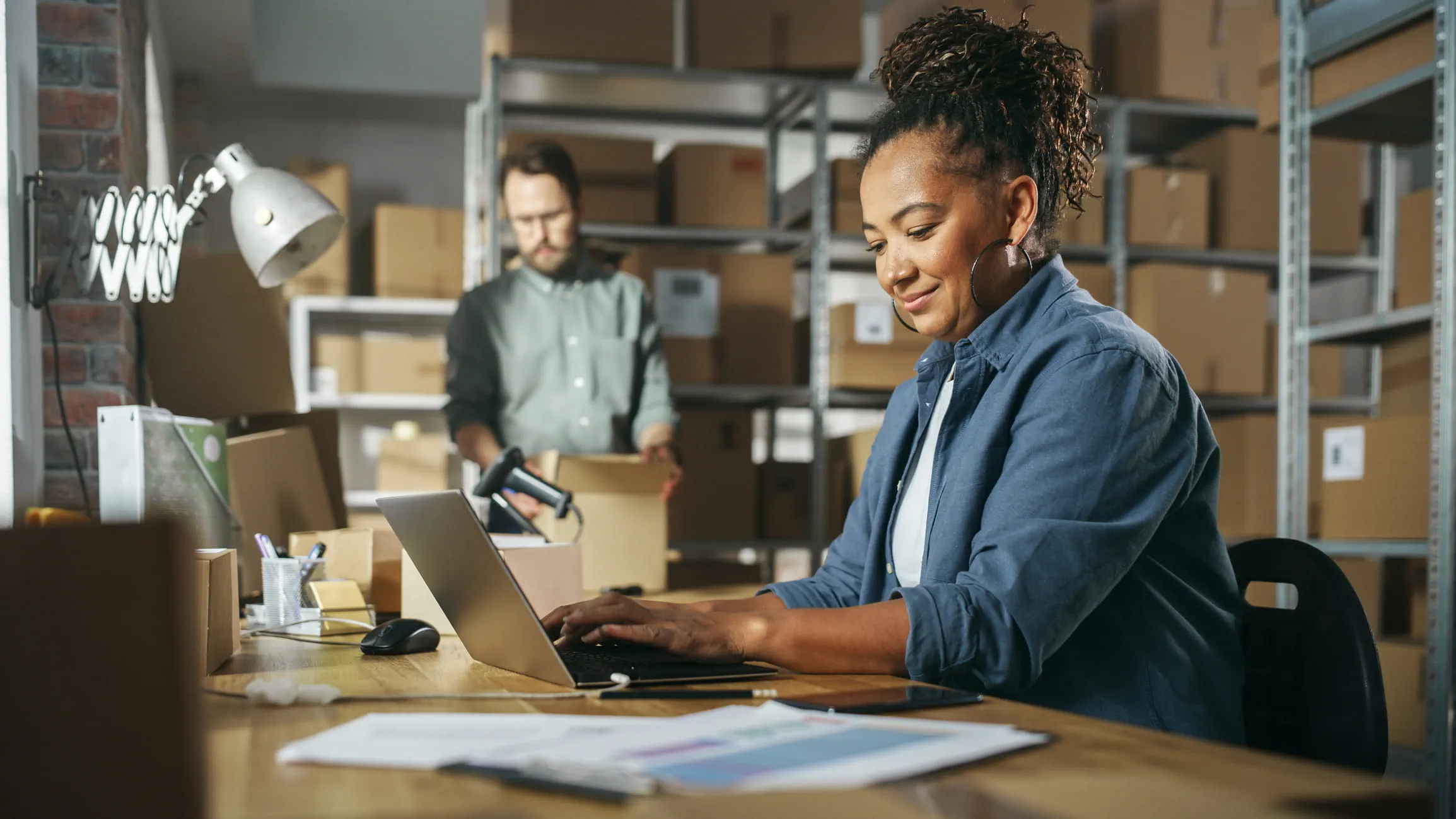 A person sits at a cluttered desk at a laptop in a shipping warehouse. A worker behind them looks at an opened box. A person sits at a cluttered desk at a laptop in a shipping warehouse. A worker behind them looks at an opened box.
