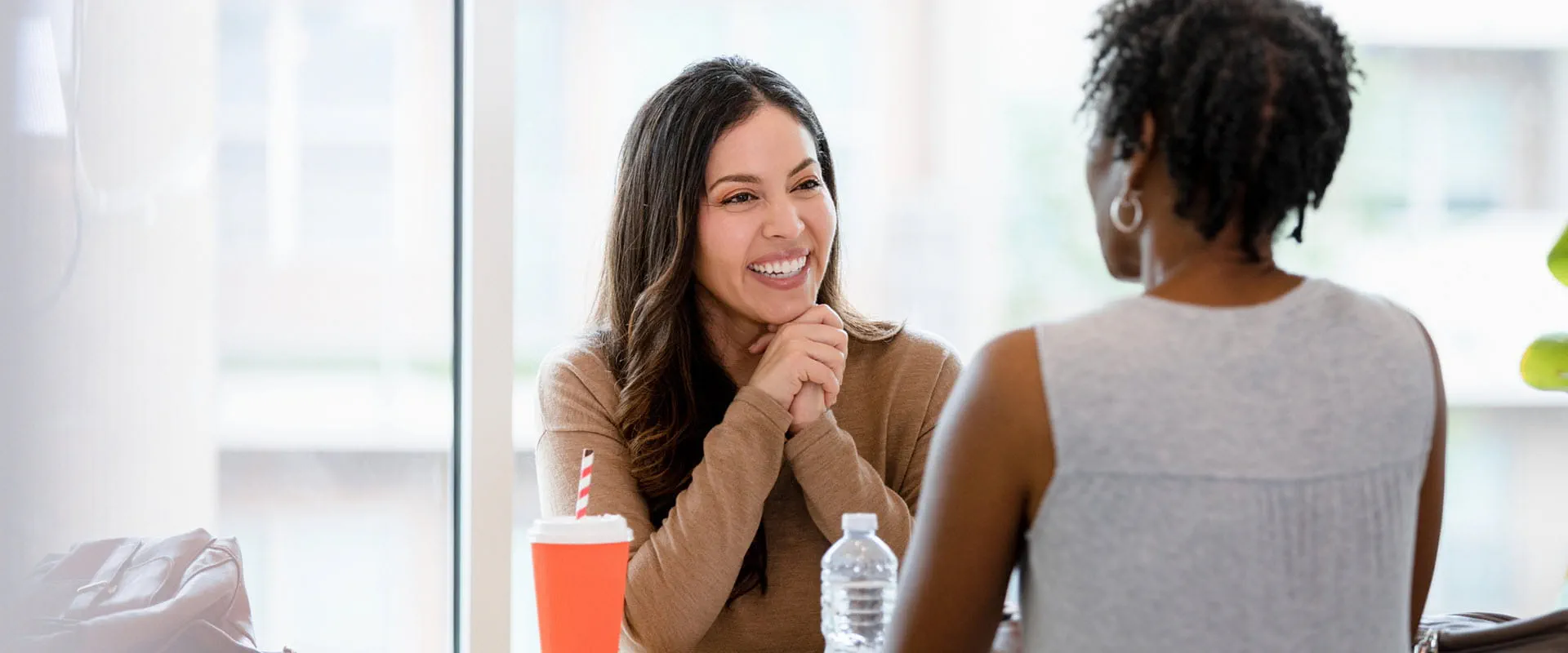 A smiling manager and employee sit across the table from one another in an office building. The table is covered in papers, coffee cup and water bottle. A smiling manager and employee sit across the table from one another in an office building. The table is covered in papers, coffee cup and water bottle.