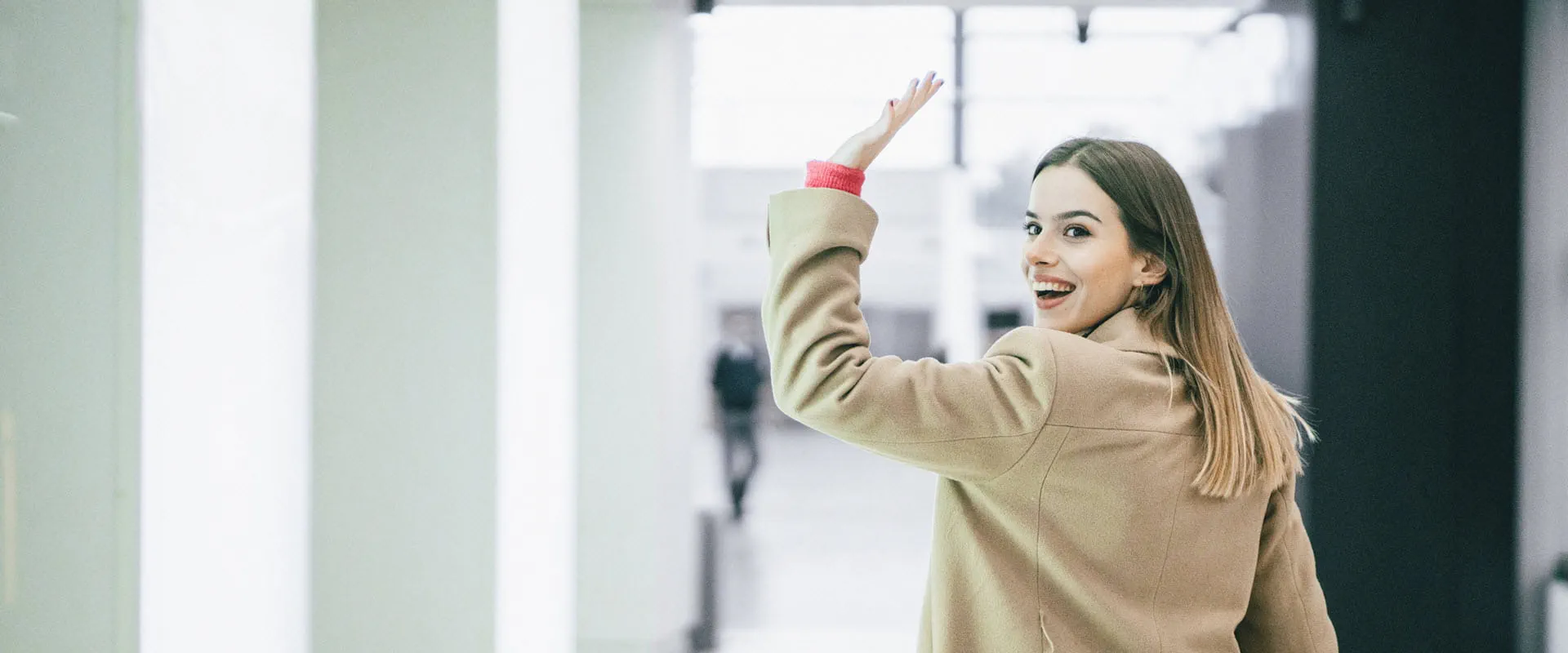 A young woman wearing a beige coat walks down a modern, well-lit hallway while waving and smiling back at the camera. A young woman wearing a beige coat walks down a modern, well-lit hallway while waving and smiling back at the camera.