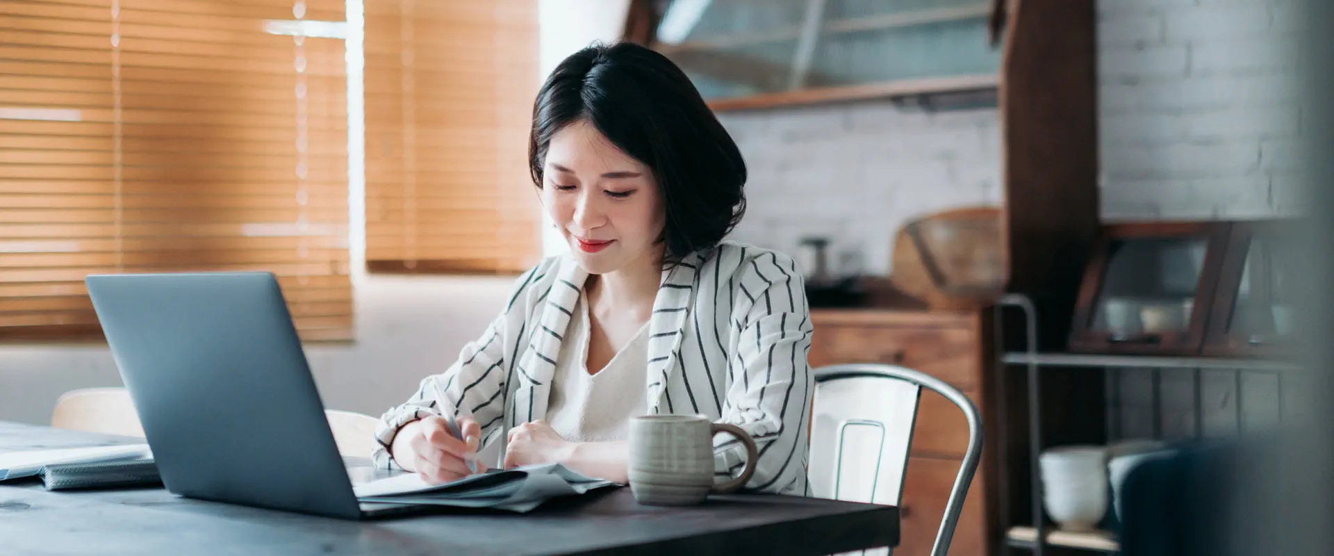 A worker sits at their kitchen table with their laptop, writing something down in a notebook. A worker sits at their kitchen table with their laptop, writing something down in a notebook.