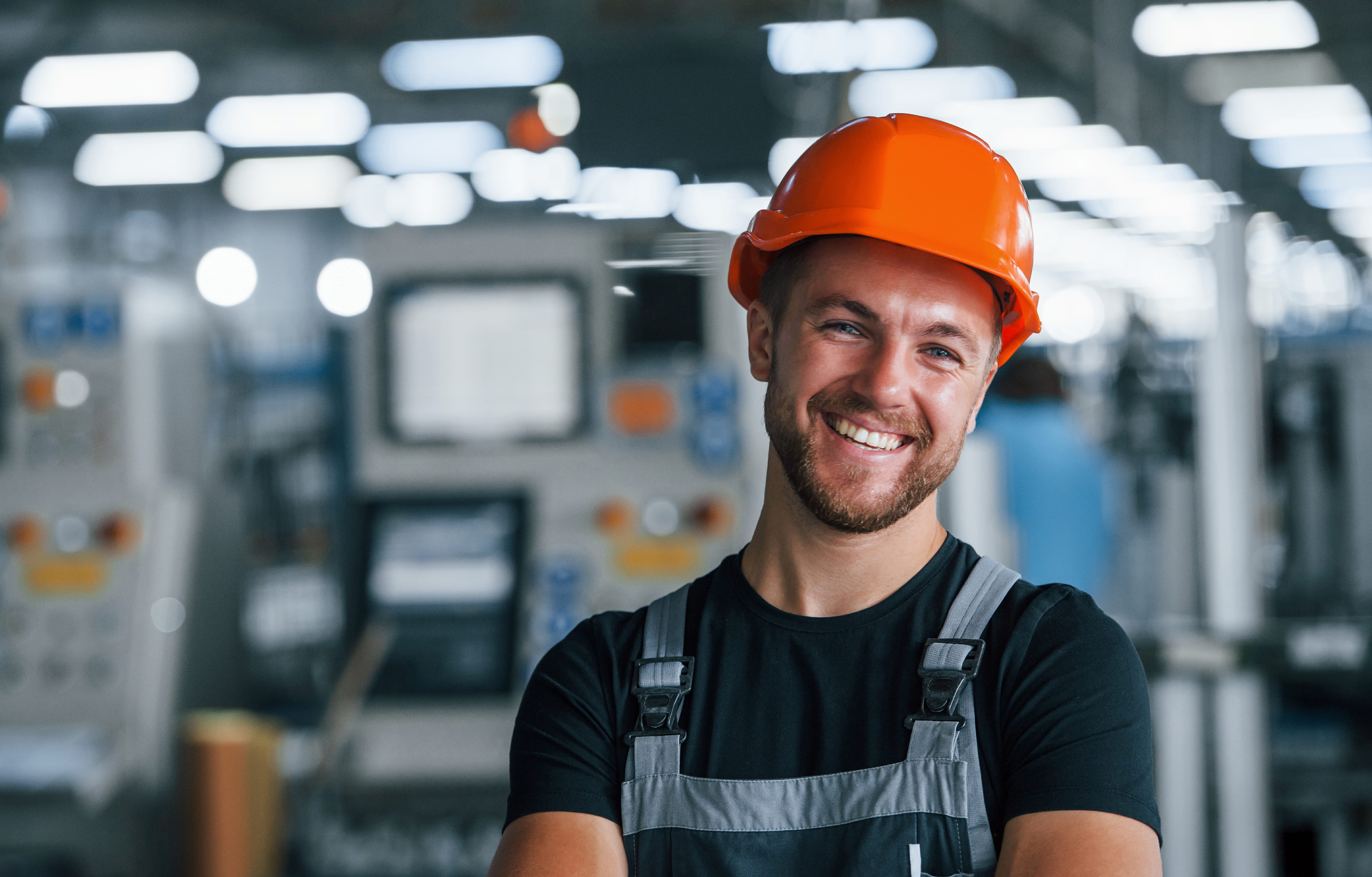 A smiling worker wearing an orange hard hat and overalls in a manufacturing facility.