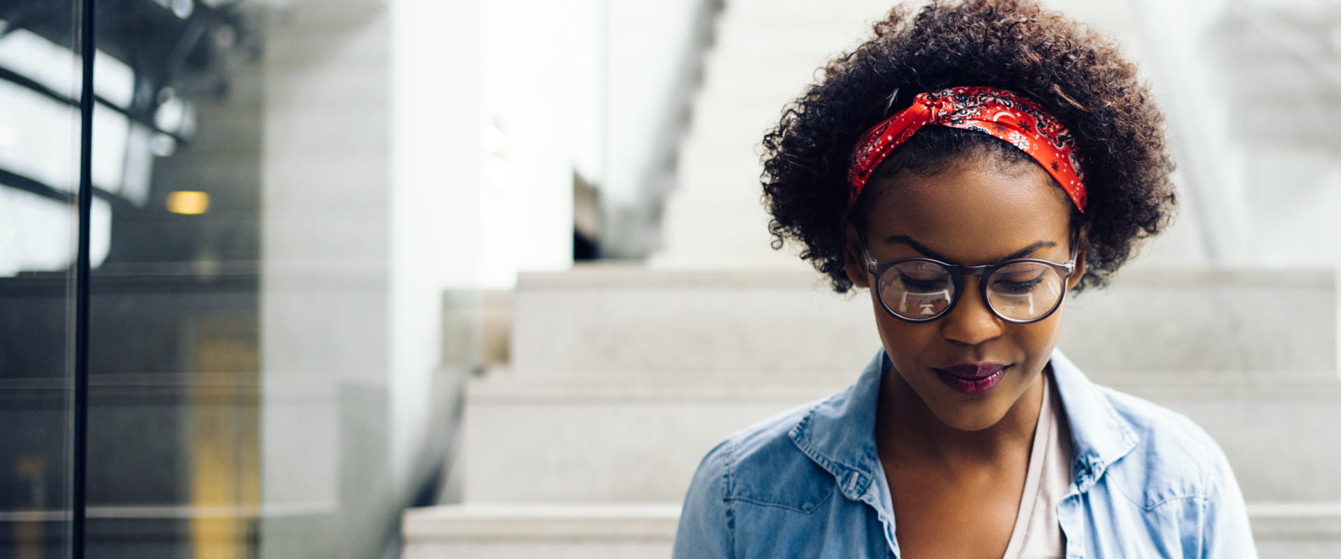 Person with glasses and a red headband looking down, standing in front of steps in an urban setting.