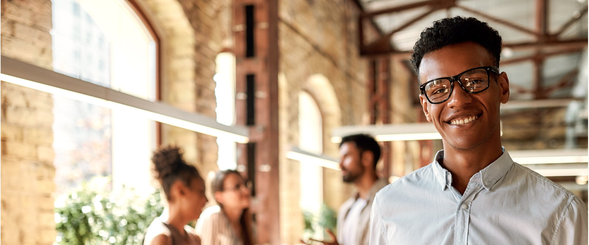 Smiling person wearing glasses in loft office space with blurred coworkers in the background.
