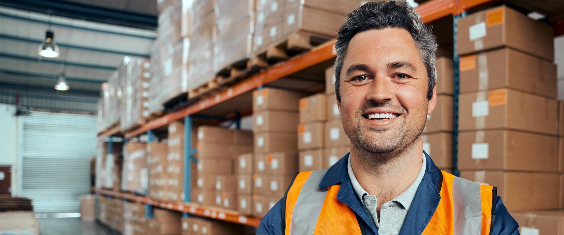 A smiling professional wearing a safety vest in a busy storage facility, with shelves of boxes in the background.