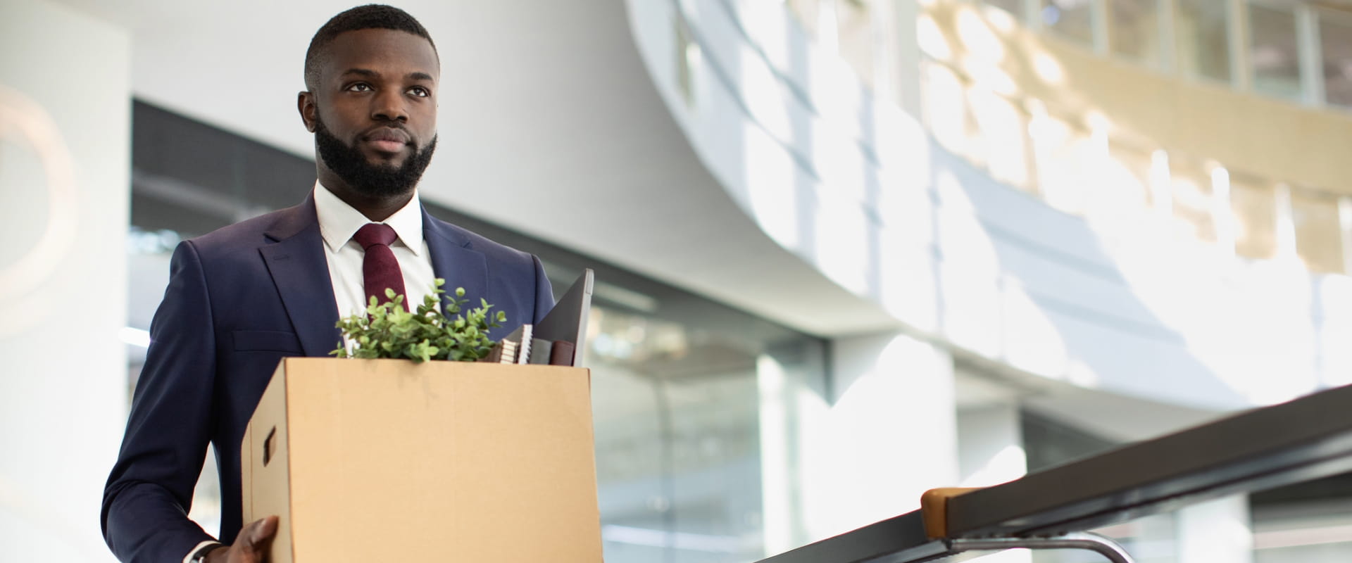 A worker in a suit seriously walks through the lobby of a large office building. He is carrying a box filled with personal items. A worker in a suit seriously walks through the lobby of a large office building. He is carrying a box filled with personal items.