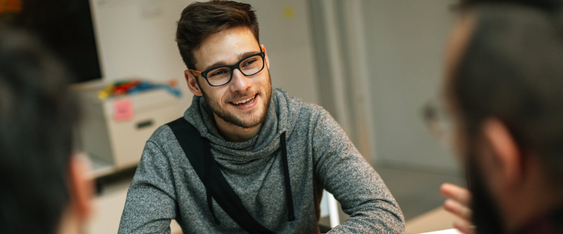 A young person with glasses, casually dressed, leans forward on a desk listening to someone. A young person with glasses, casually dressed, leans forward on a desk listening to someone.