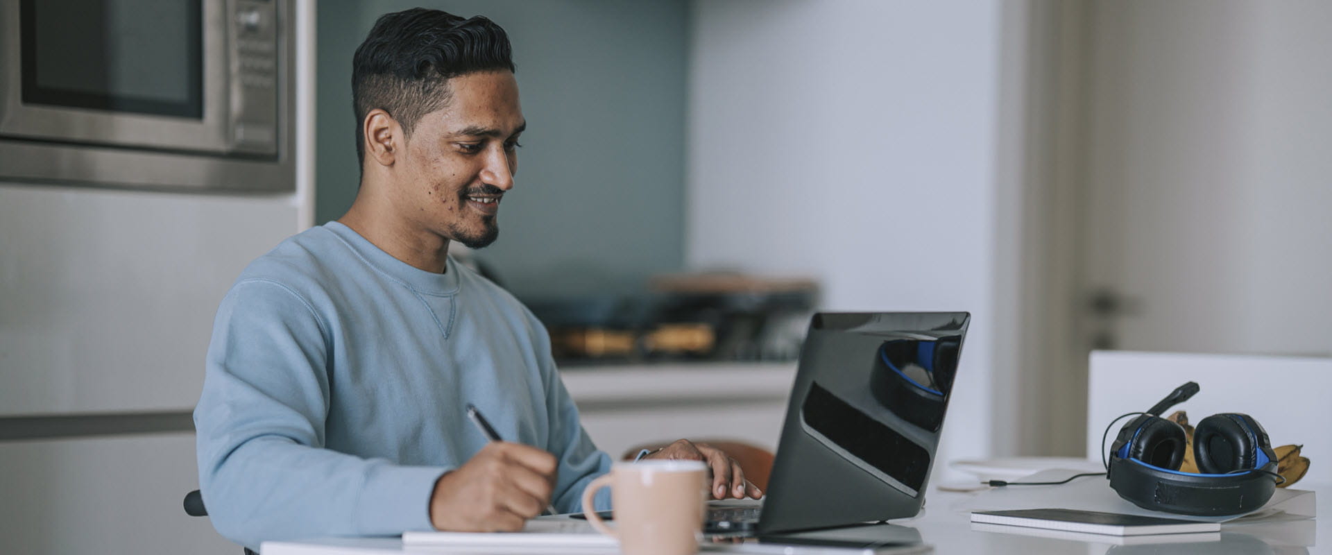 A person in a wheelchair is at their kitchen table, writing something in a notepad while looking at their laptop. A person in a wheelchair is at their kitchen table, writing something in a notepad while looking at their laptop.